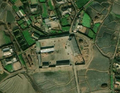 4/6 Cour de ferme (landuse=farmyard) composée de longs bâtiments entouré d'une clôture parsemée des tas de foins, à proximité d'une route très proche d'un village (imagerie satellite Maxar).
