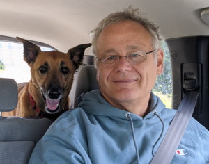 Picture of Craig Allan in the driver's seat of a car, smiling, in Montana, with a curious dog.